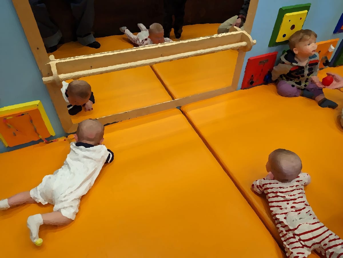 Two babies on their bellies in front of a mirror stare at their reflections